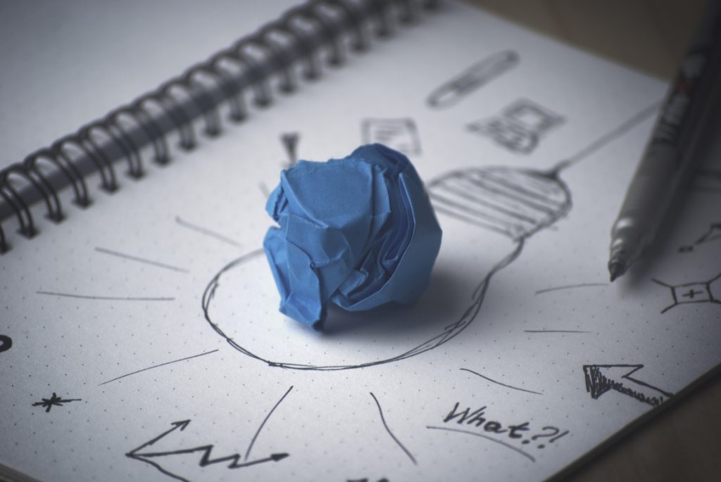 Image to represent critical thinking and the cross-section with creativity. A sketch pad with a drawing of a light bulb. There are various lines coming leading out from the light bulb. The word What? is prominent in the foreground. A pen rests on the page. A blue crumpled ball of paper is place in the center of the light bulb.